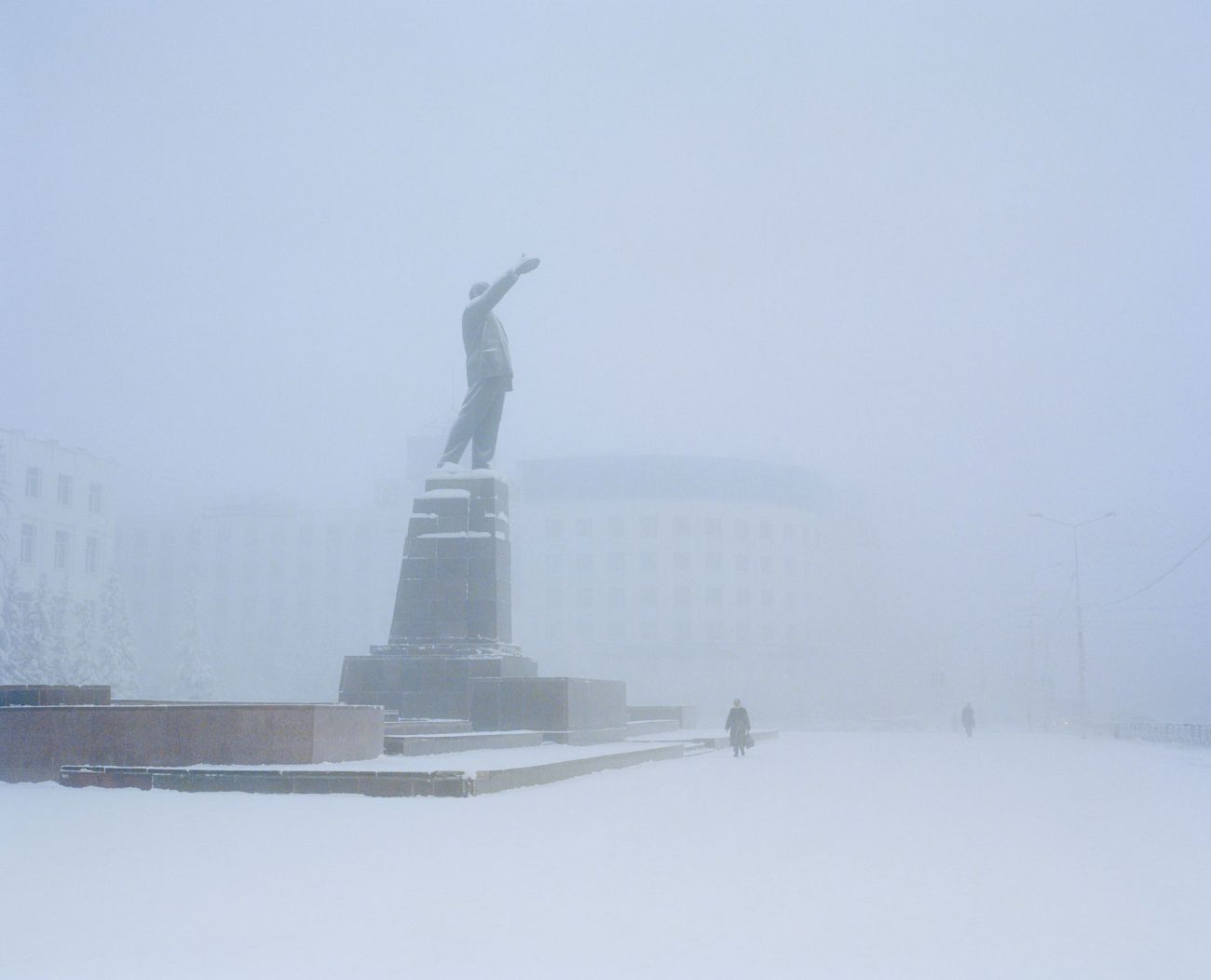 Sacha by Alexis Pazoumian — A bronze statue of a figure with raised arm stands on a monumental pedestal within a snow-covered public square in Yakutsk.