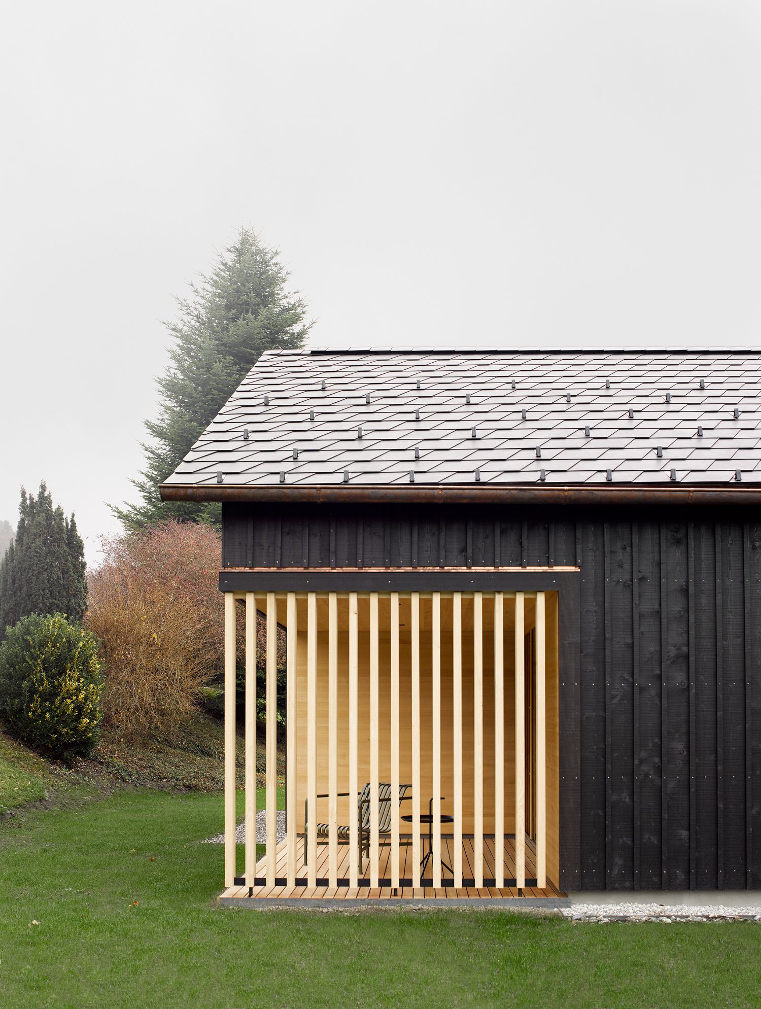 House ZF by Bernd Riegger — A timber-framed garden pavilion extends from the dark-clad main structure, its cream-painted vertical slats creating a semi-permeable threshold between...