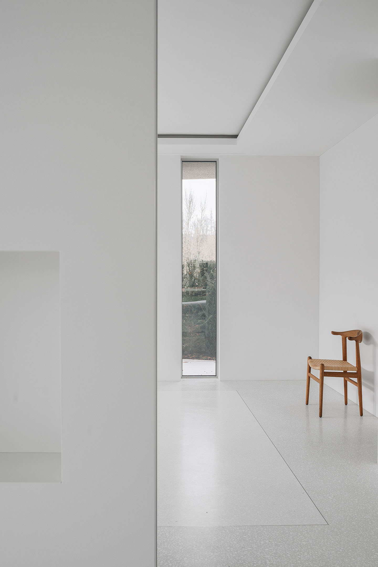 House for an Art Collector by Think Architecture — A minimalist interior passage demonstrates material hierarchy through a narrow vertical window framing bare winter landscape.