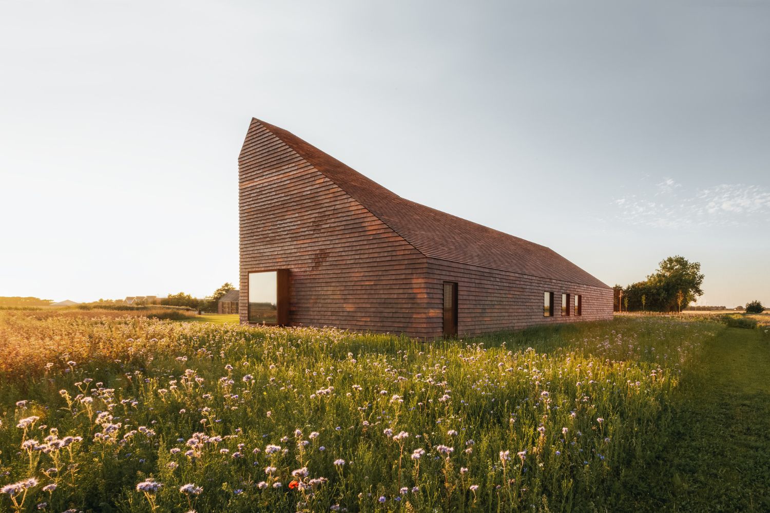 House of Grain by Reiulf Ramstad Arkitekter — The grain house reveals its volumetric brick form against a cultivated field dotted with wildflowers and grasses.