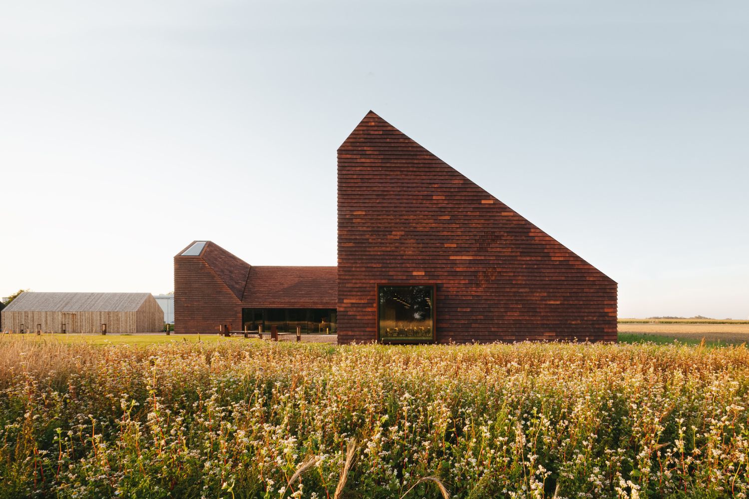 House of Grain by Reiulf Ramstad Arkitekter — The House of Grain rises from a mature grain field in golden-hour light, its composition anchored by two pyramidal brick towers with steeply pitched roofs.