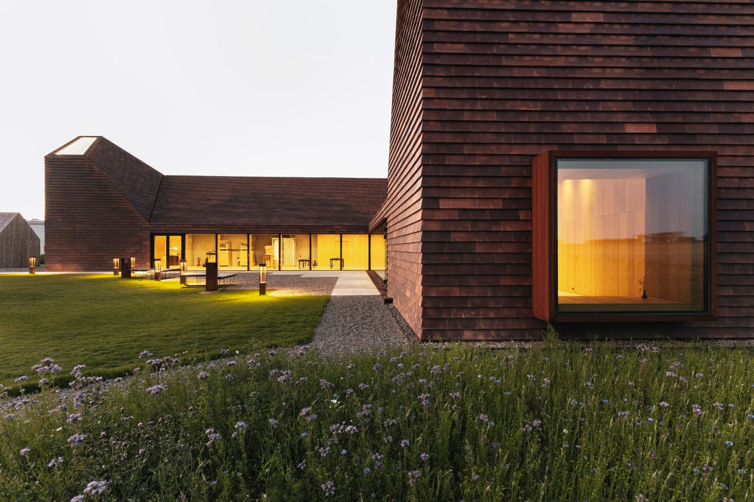 House of Grain by Reiulf Ramstad Arkitekter — Twilight view of the House of Grain's eastern facade, featuring a large rectangular dark-framed window portal with golden interior light flooding the...