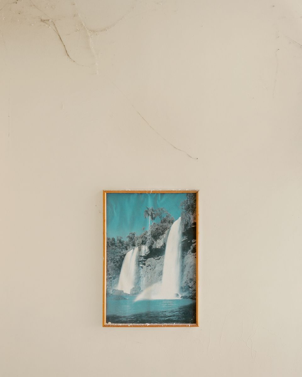 Along the Swift Current of Time by Teodora Georgieva — A framed photograph of twin waterfalls cascades into turquoise water, mounted within a weathered timber frame against a deteriorating cream plaster wall.