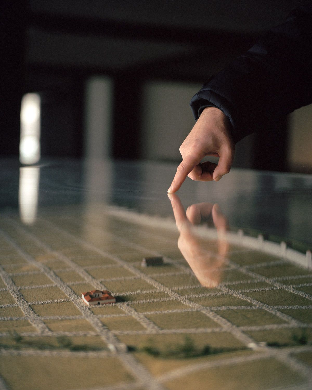 Along the Swift Current of Time by Teodora Georgieva — A hand in dark fabric hovers above a miniature landscape model, index finger extended in a gesture of selection or intervention.