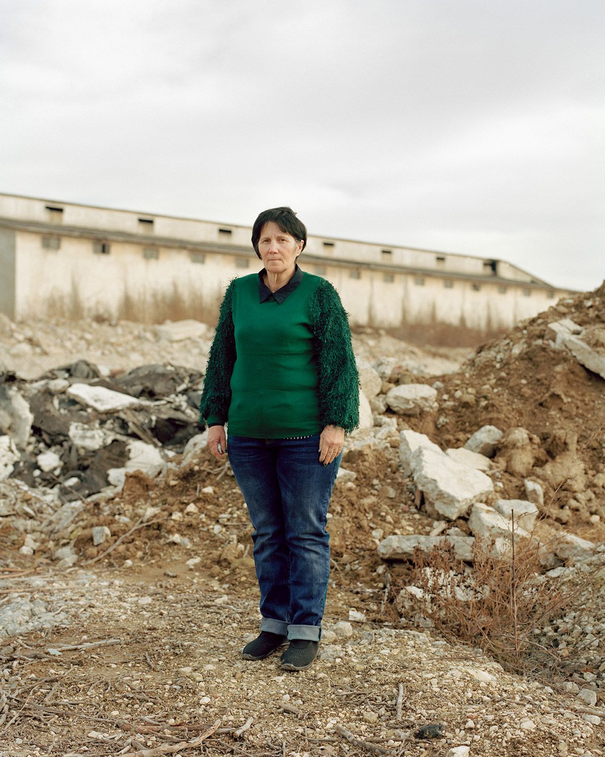 Along the Swift Current of Time by Teodora Georgieva — A woman stands centered in a barren industrial landscape, wearing a green textured cardigan over dark jeans.