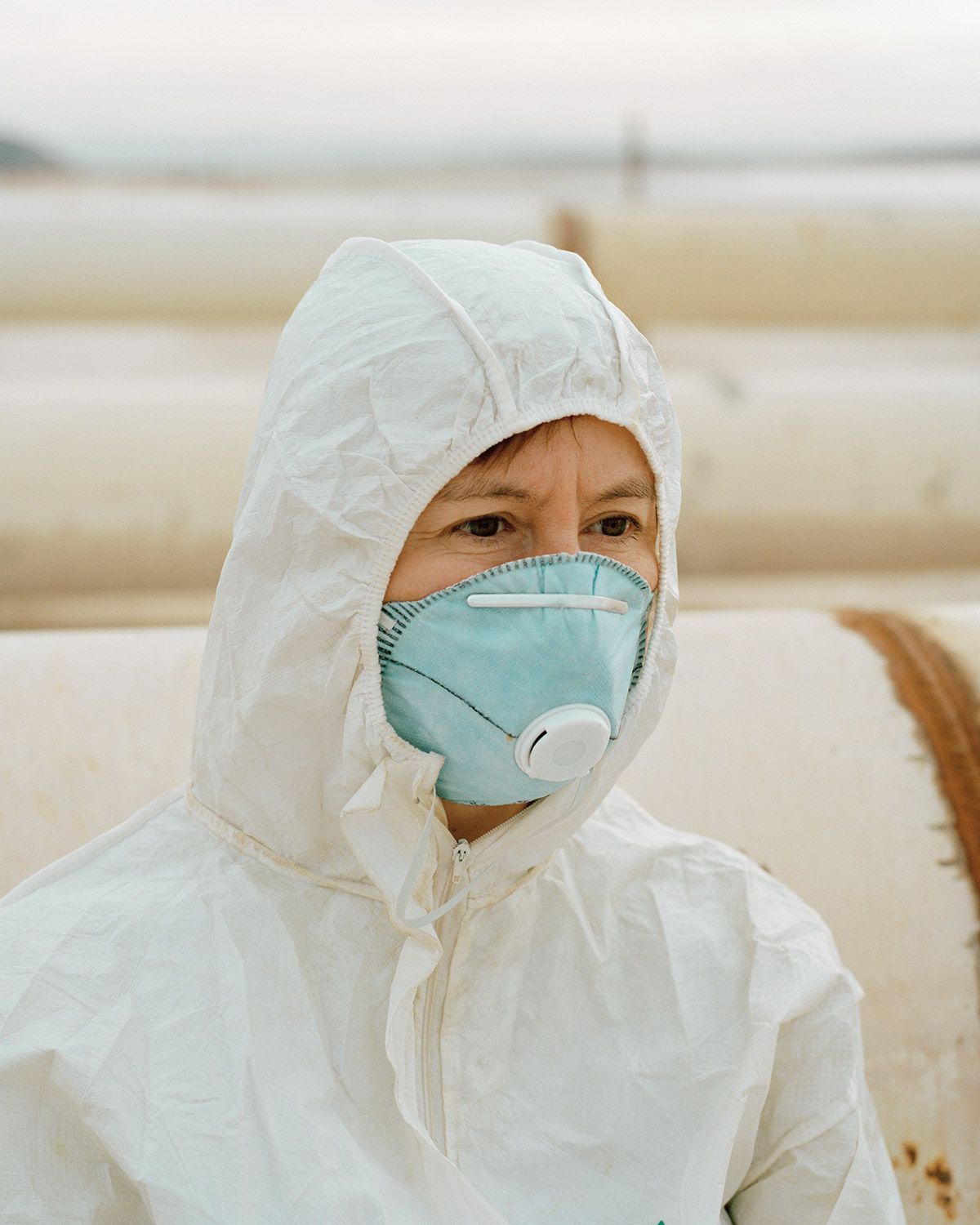 Along the Swift Current of Time by Teodora Georgieva — A child in protective white coveralls and a turquoise respirator mask stands against an industrial backdrop of pale mineral deposits and layered sediment...