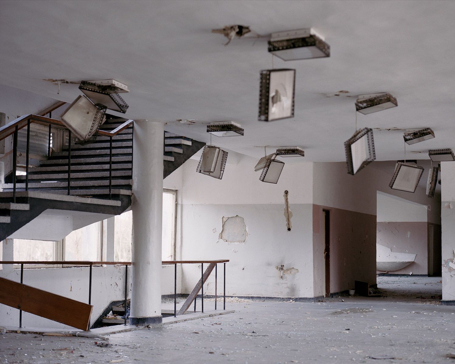 Along the Swift Current of Time by Teodora Georgieva — An abandoned institutional interior features suspended film reels or projection equipment mounted across a white ceiling, their metal frames casting...