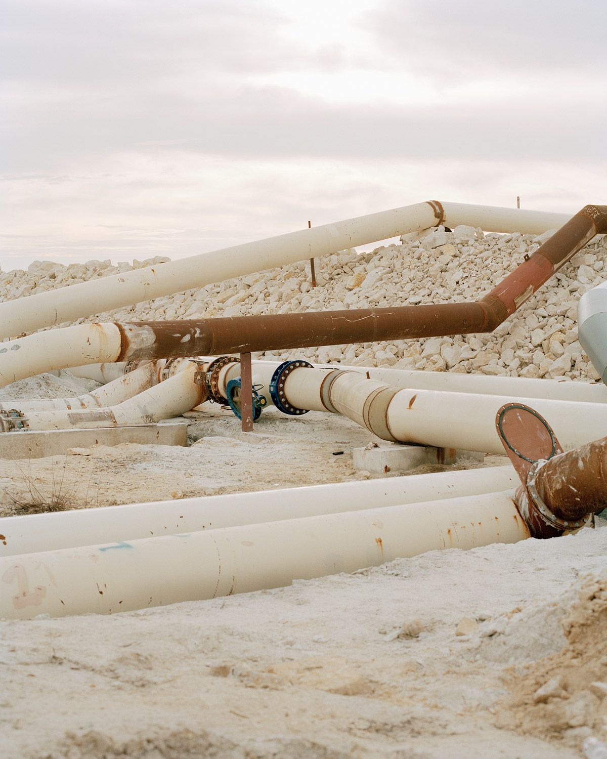 Along the Swift Current of Time by Teodora Georgieva — Industrial pipes in various stages of deterioration lay across a barren landscape scattered with white mineral deposits and aggregate material.