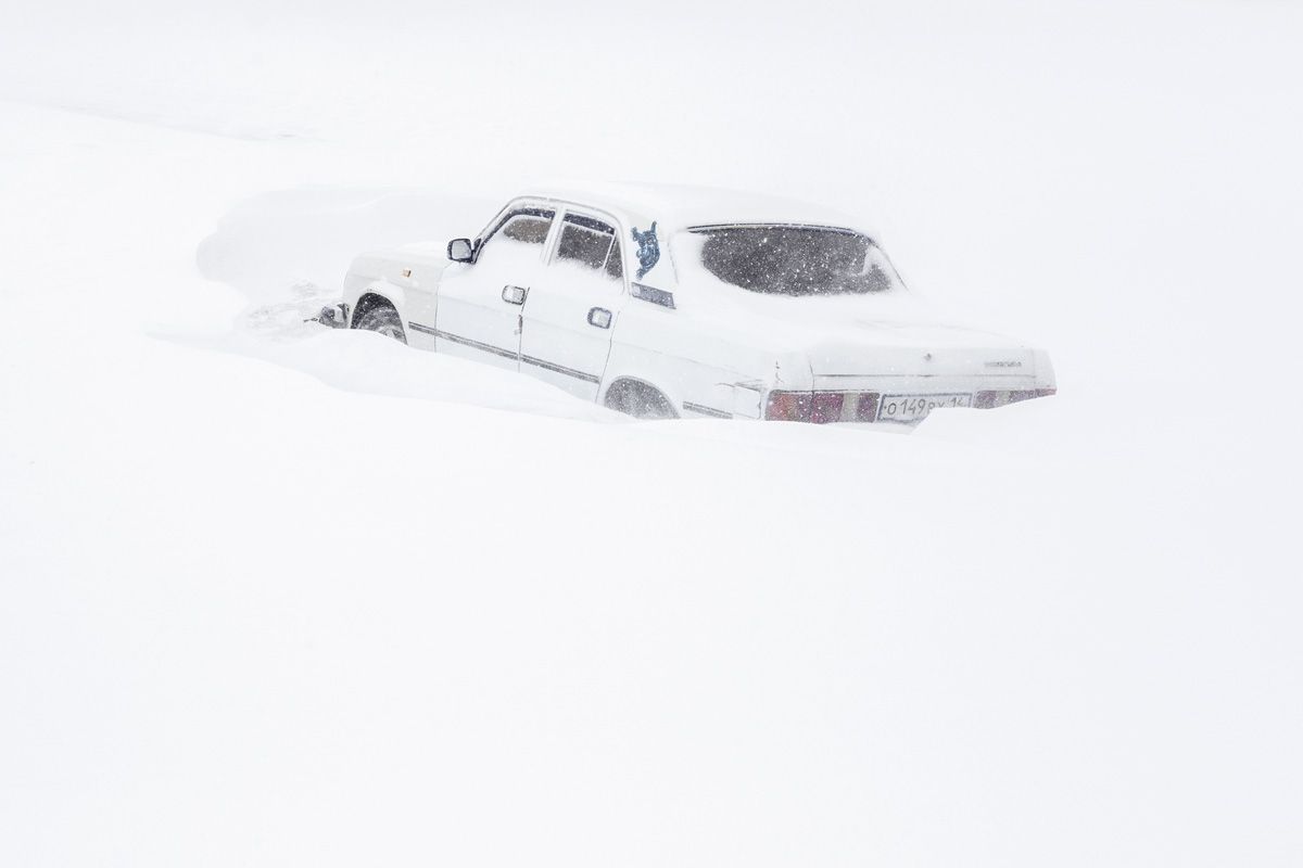 Удачный / Lucky by Hanne Van Assche — A white sedan lies half-submerged in deep snow, its elongated body stretched horizontally across a monochromatic landscape.