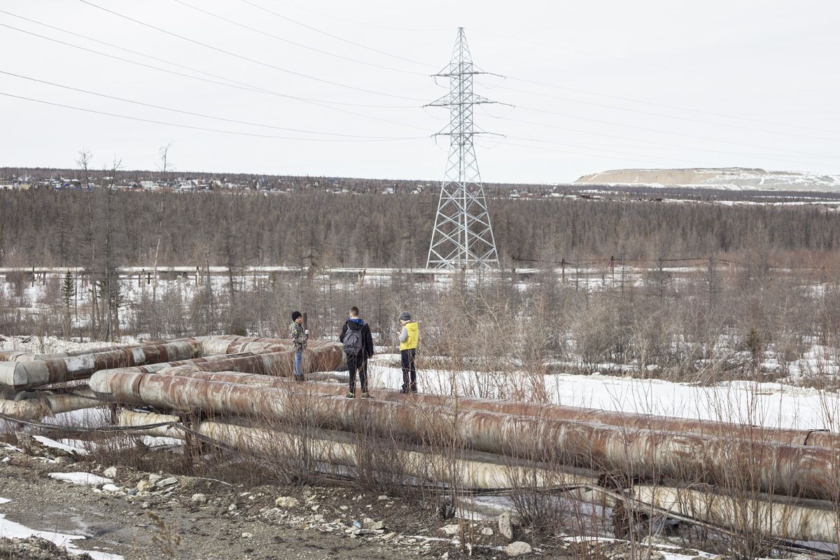 Удачный / Lucky by Hanne Van Assche — Three figures stand among rusted industrial pipes in a snow-patched clearing, with a high-voltage transmission tower dominating the composition behind them.