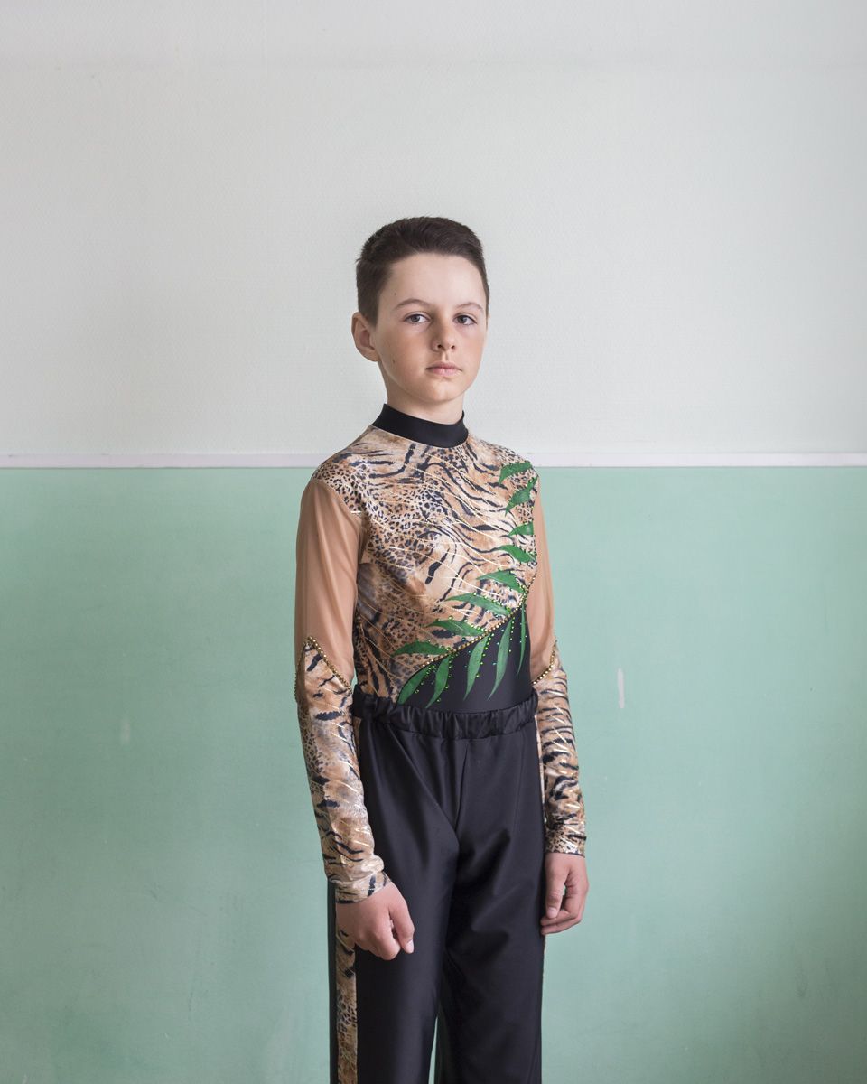 Удачный / Lucky by Hanne Van Assche — A young boy stands in studio against a horizontally divided backdrop of white upper section and sage lower section.