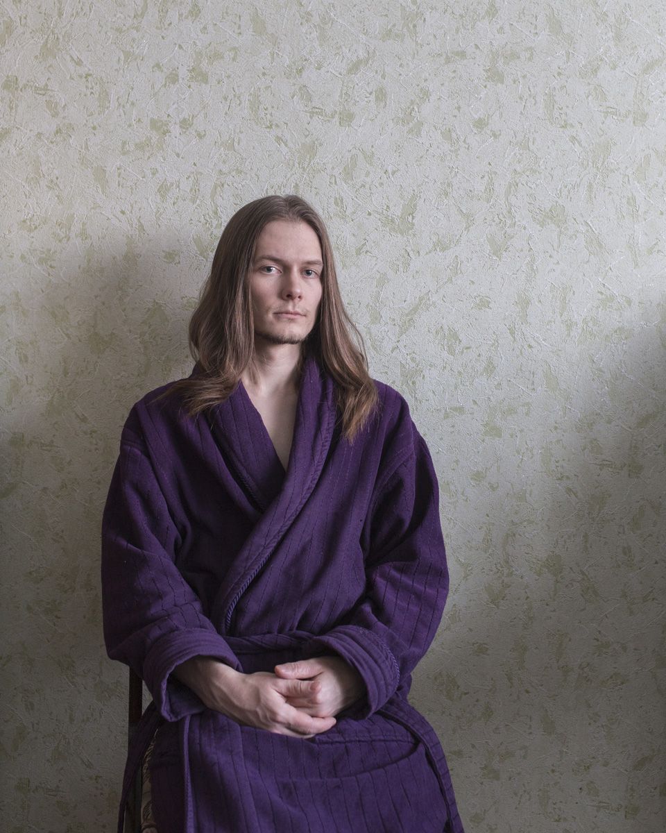 Удачный / Lucky by Hanne Van Assche — A portrait photograph of a person seated against a textured plaster wall in pale grey-green tones.