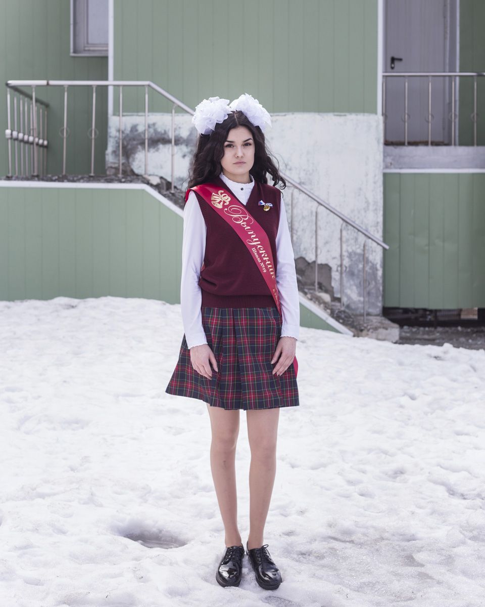 Удачный / Lucky by Hanne Van Assche — A young woman stands on snow in the mining town of Udachny, wearing a burgundy tartan skirt, maroon knit vest with embroidered lettering, white long-sleeved...