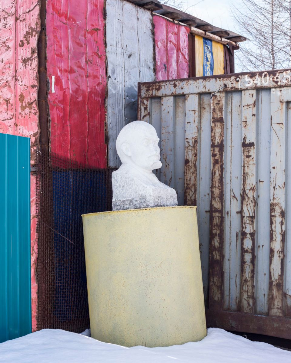 Удачный / Lucky by Hanne Van Assche — A white plaster bust emerges from a yellow cylindrical vessel positioned against weathered industrial structures.