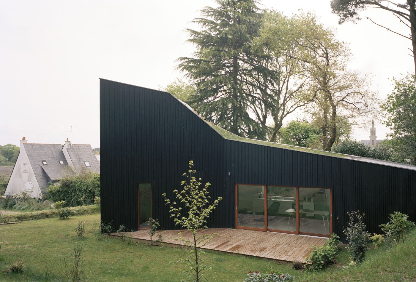 Holiday House in Sarzeau by RAUM — A dark corrugated metal structure with a dramatically pitched roof dominates the foreground, its angular profile rising above mature cedar and pine trees.