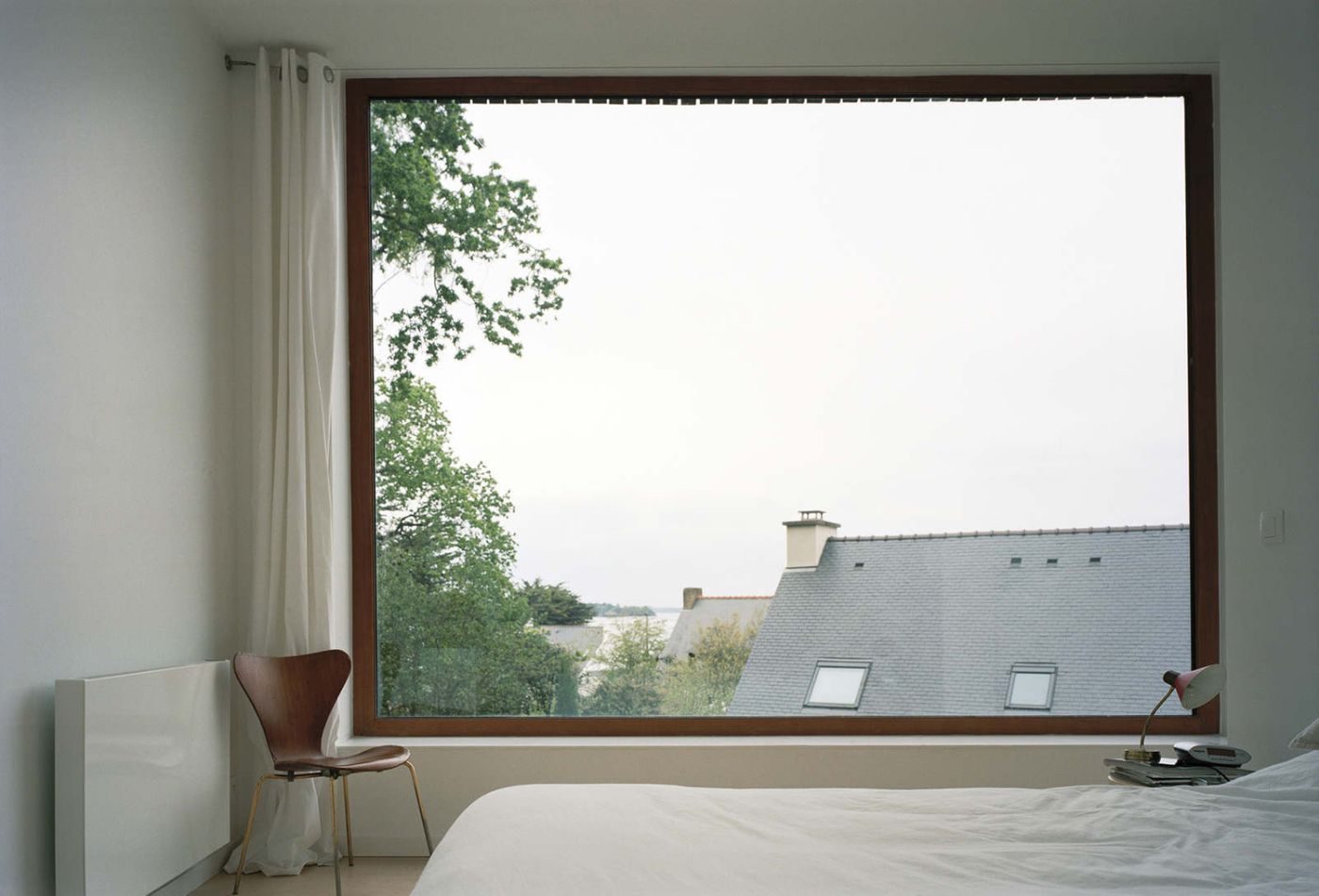 Holiday House in Sarzeau by RAUM — A bedroom interior frames an expansive horizontal window revealing slate-roofed buildings and mature trees beyond.