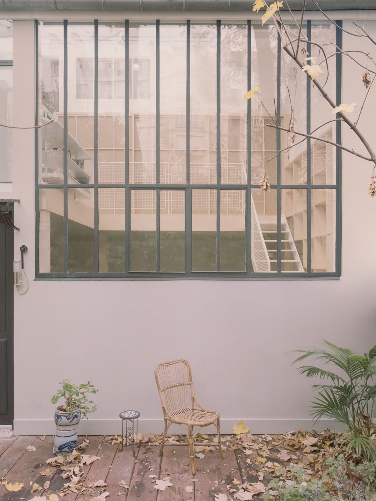 Jean Moulin Atelier – House by Atelier NEA — A sheltered courtyard garden with a large steel-framed window system overlooking an adjacent building's staircase and facade.