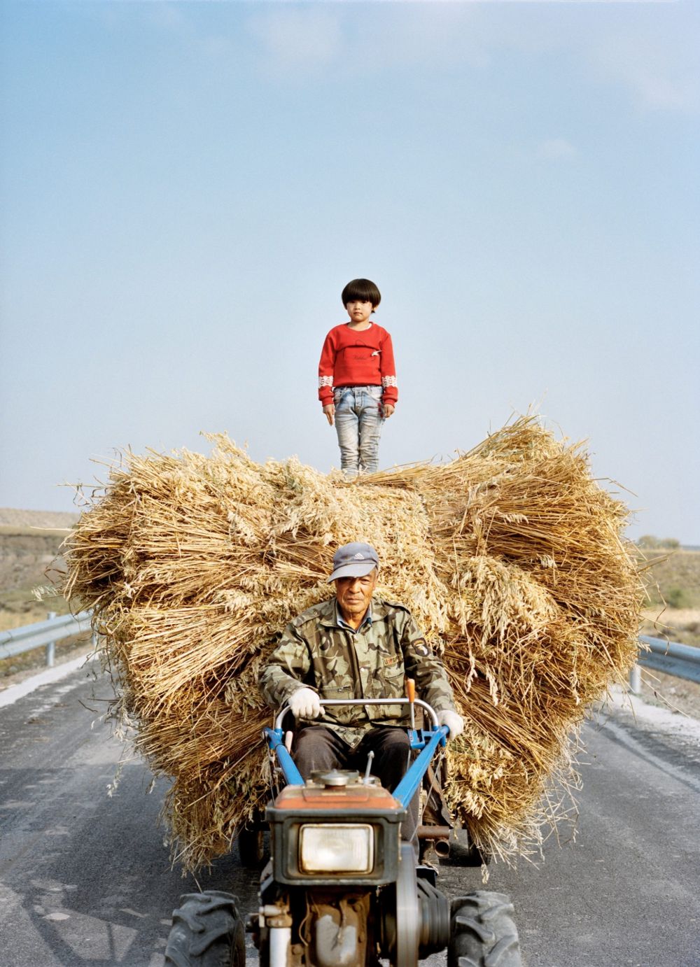 Watering My Horse by Xiaoxiao Xu — A man operating a motorized three-wheeler laden with an oversized bundle of dried straw navigates a paved road through rural terrain near the Great Wall.