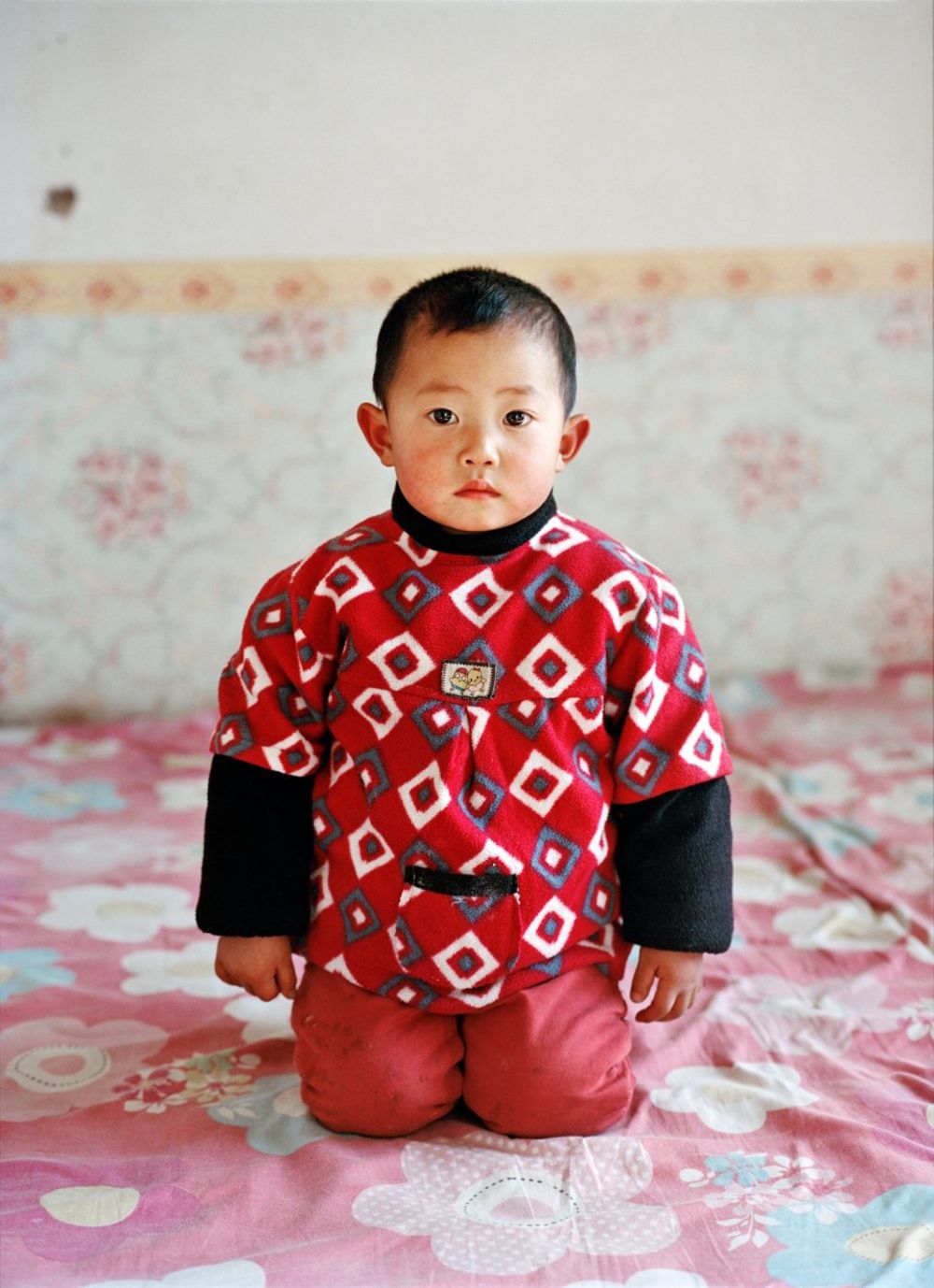 Watering My Horse by Xiaoxiao Xu — A young child in a red geometric-patterned sweater with black long sleeves kneels on a pink floral bedspread, positioned centrally against a wallpapered...