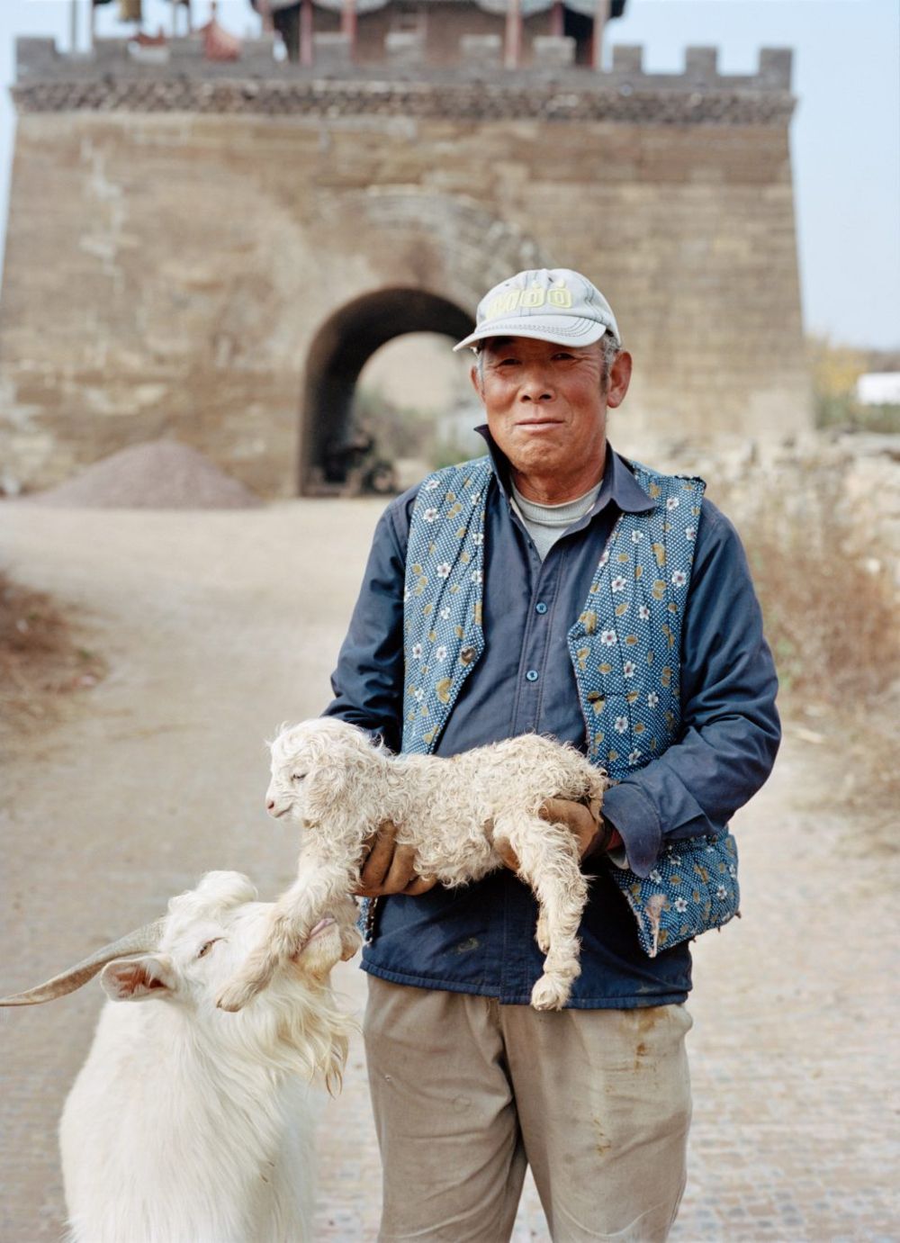 Watering My Horse by Xiaoxiao Xu — A man in a white baseball cap and layered blue jacket with geometric patterning holds a young kid goat while standing beside an adult white goat on a dirt path.