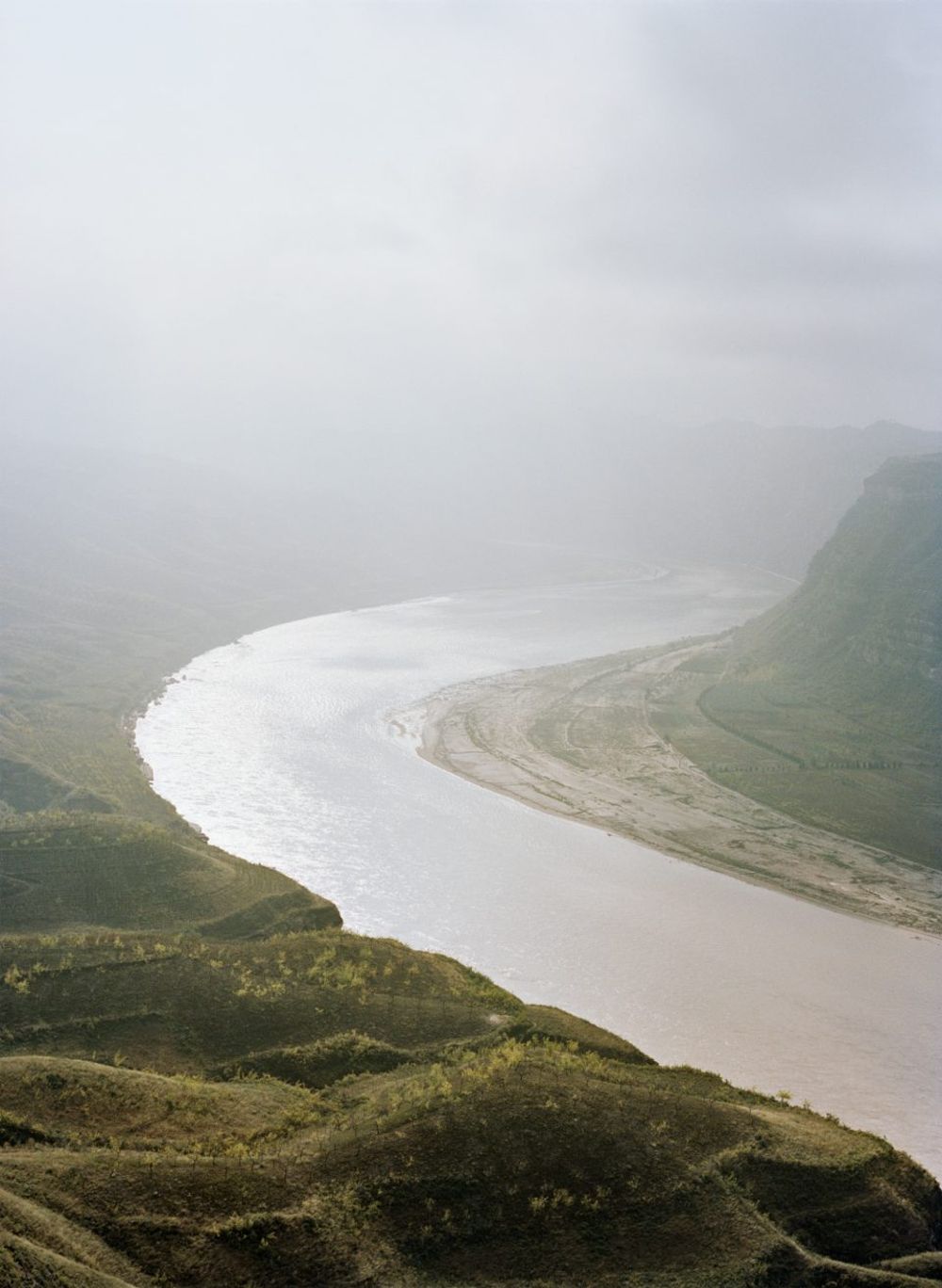 Watering My Horse by Xiaoxiao Xu — A serpentine river winds through a valley beneath overcast skies, its pale silted waters contrasting sharply against exposed earth banks and sparse vegetation.