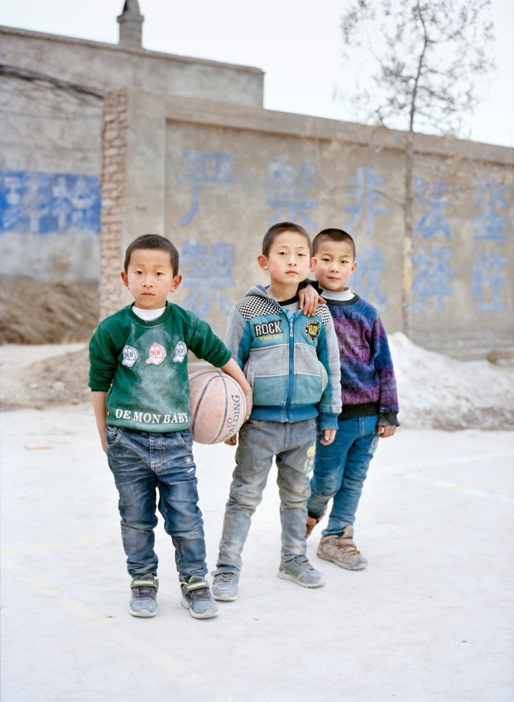 Watering My Horse by Xiaoxiao Xu — Three boys stand in formation on packed earth before a section of Ming dynasty stone wall, its rectangular blocks marked with faded blue characters.