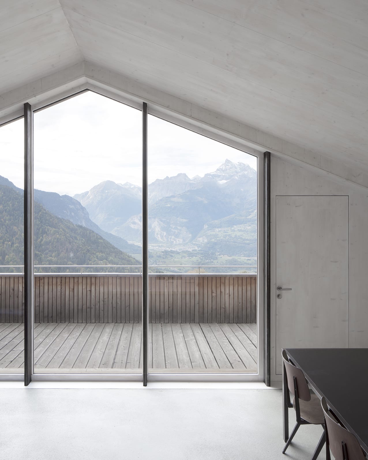 En Chalméry House by Christian Dupraz Architects — A threshold framed by floor-to-ceiling glazed doors with dark steel mullions opens onto a timber-decked terrace overlooking an Alpine valley.