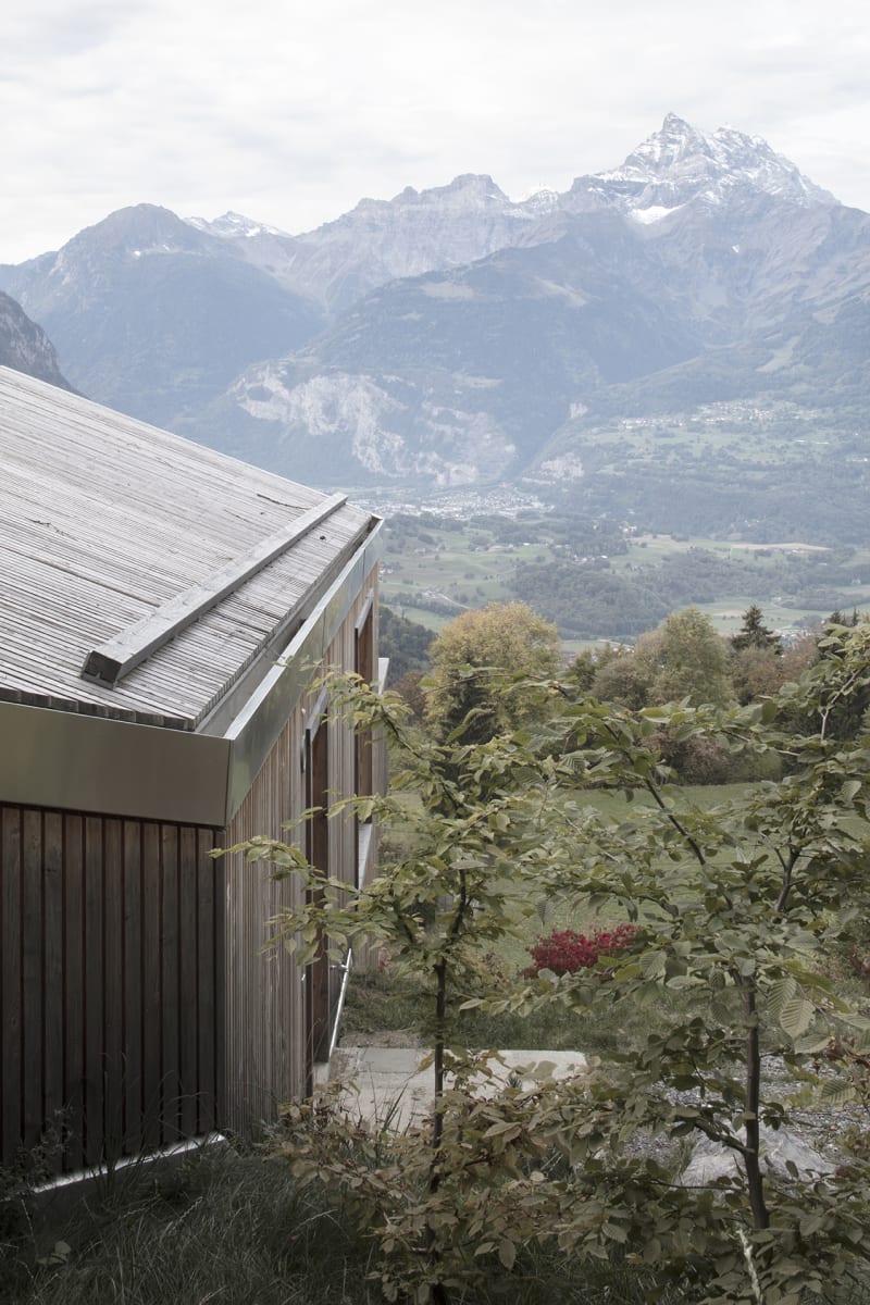 En Chalméry House by Christian Dupraz Architects — A modern timber-clad structure anchors the foreground of this Alpine landscape photograph, its dark corrugated metal siding and pitched metal roof creating...