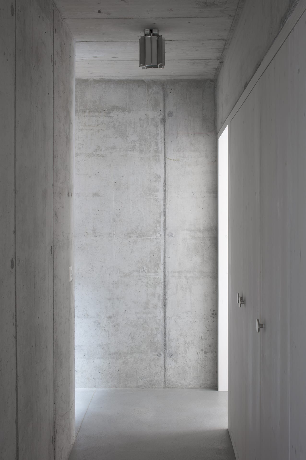 En Chalméry House by Christian Dupraz Architects — A narrow concrete corridor in board-formed cast concrete extends toward a glazed threshold flooded with exterior light.