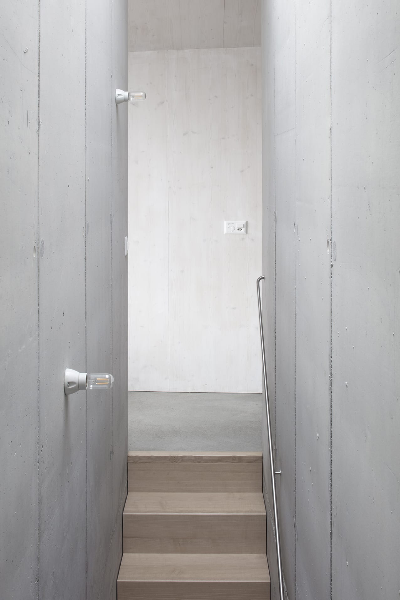 En Chalméry House by Christian Dupraz Architects — A narrow stairwell passage carved through concrete walls ascends toward diffuse light.