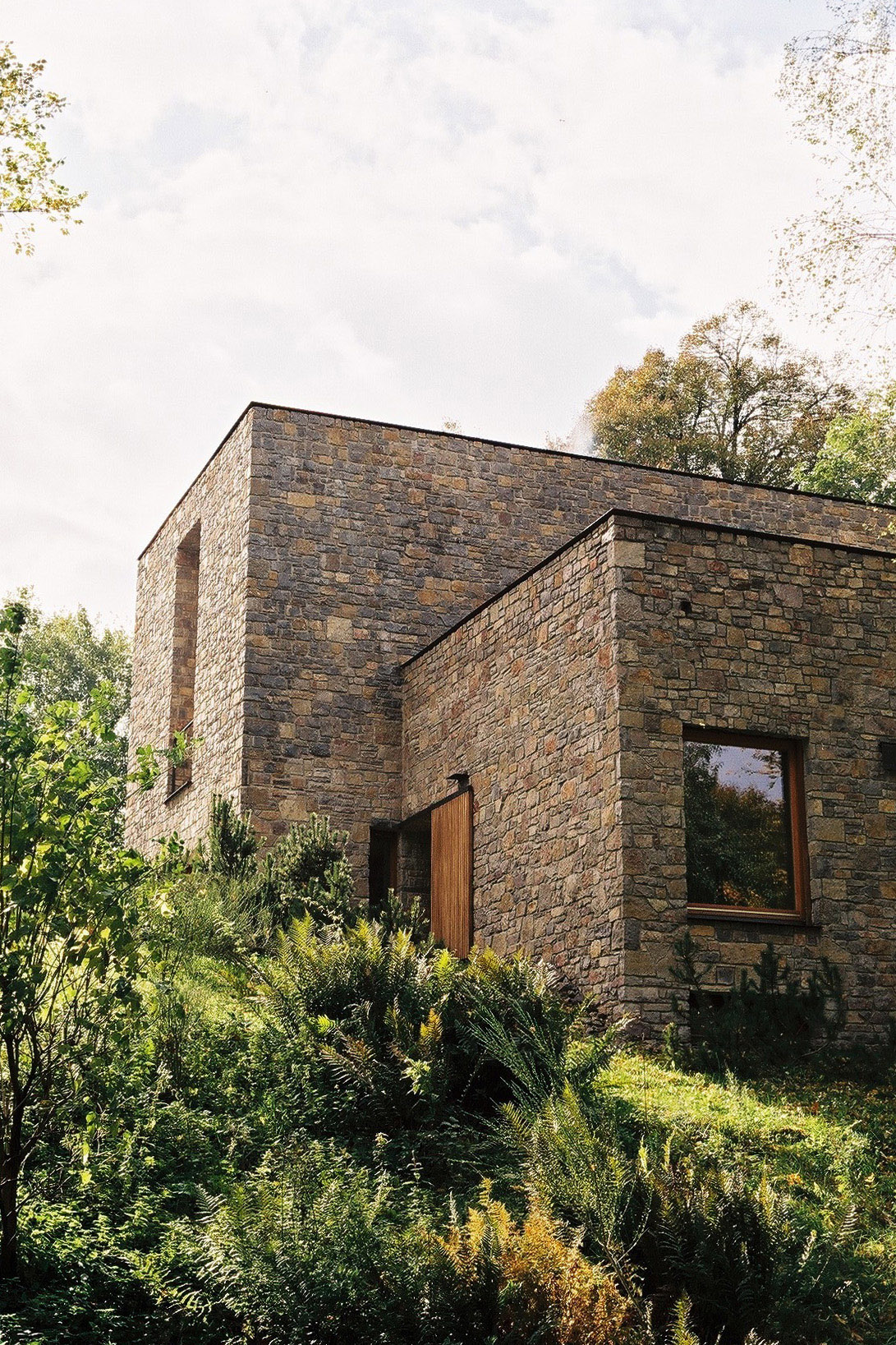 Wygnanowice House Over Crystal Clear Springs captured by Thisispaper Studio — A cubic stone tower rises above a single-story wing clad in coursed dolomite masonry, its warm ochre and grey tones darkened by weathering.