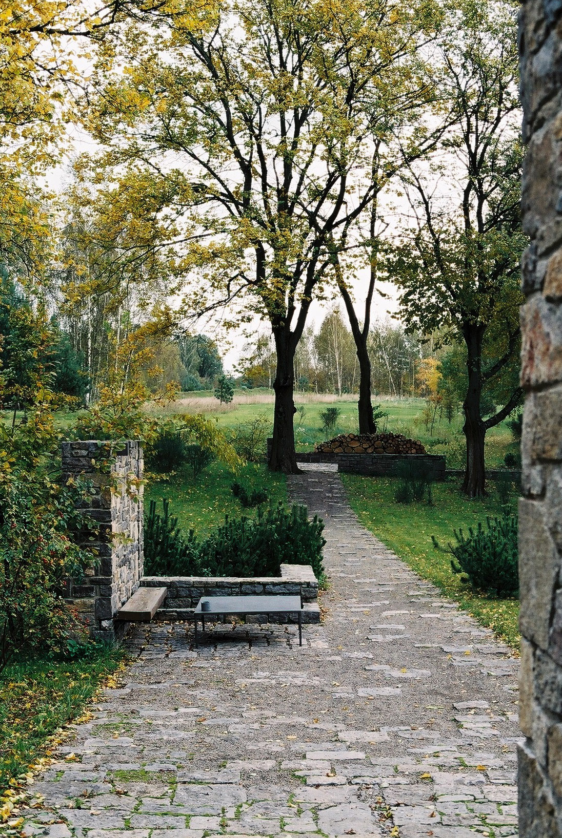 Wygnanowice House Over Crystal Clear Springs captured by Thisispaper Studio — A coursed stone pathway of grey dolomite leads through a garden threshold framed by stone masonry pillars on either side.