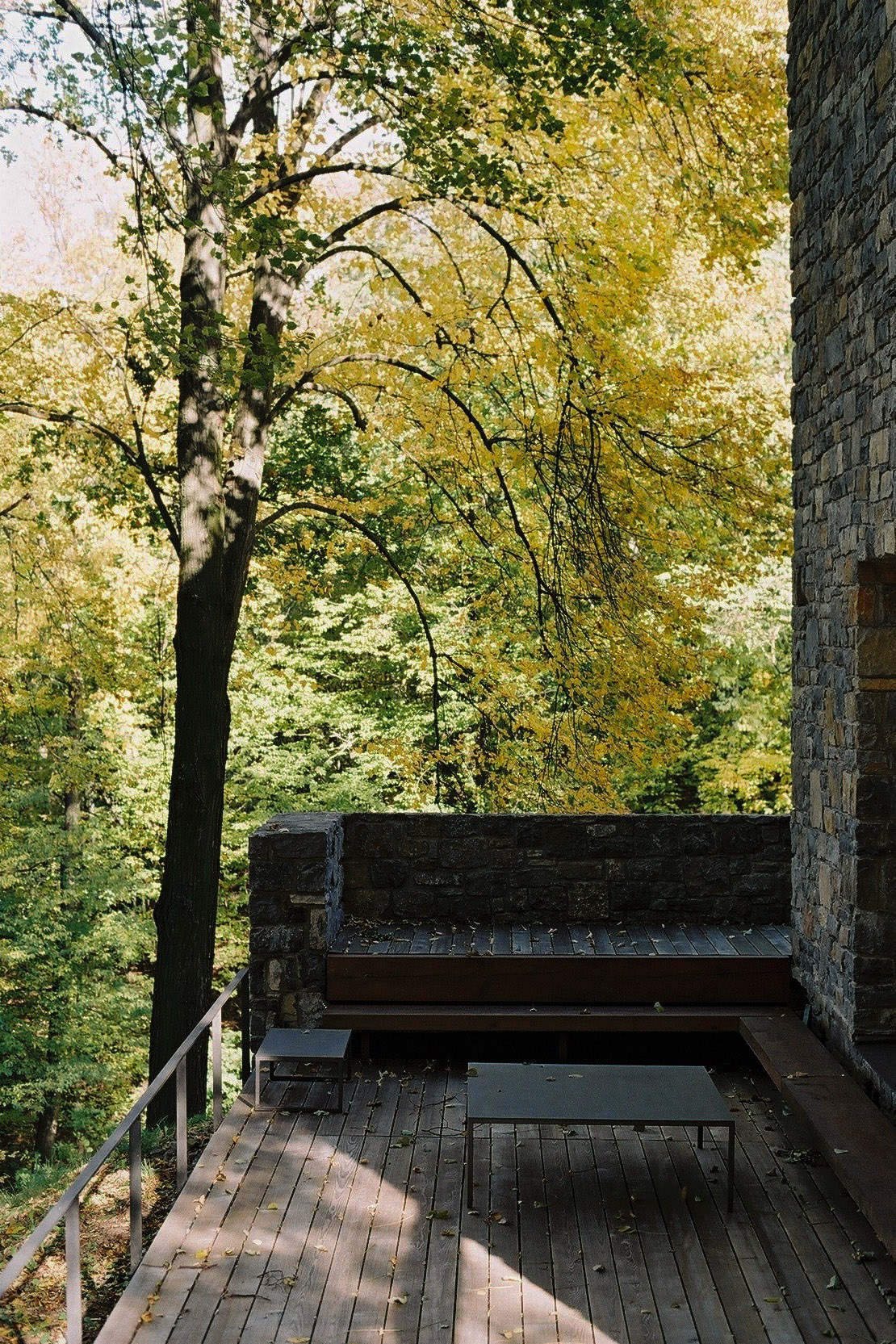 Wygnanowice House Over Crystal Clear Springs captured by Thisispaper Studio — A wooden deck extends from the stone tower, framing a view into dense autumn foliage.
