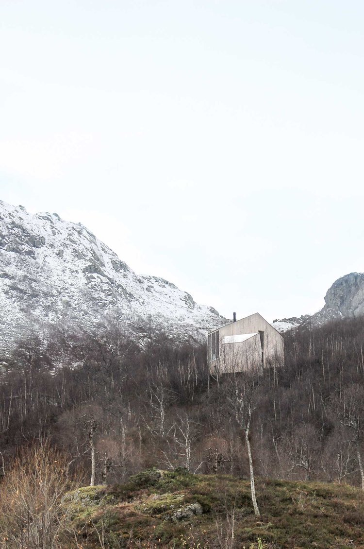Røldal Cabin by Reiulf Ramstad Arkitekter — The Røldal Cabin's upper volume is framed here against snow-capped peaks and a dense forest of bare deciduous and coniferous trees.