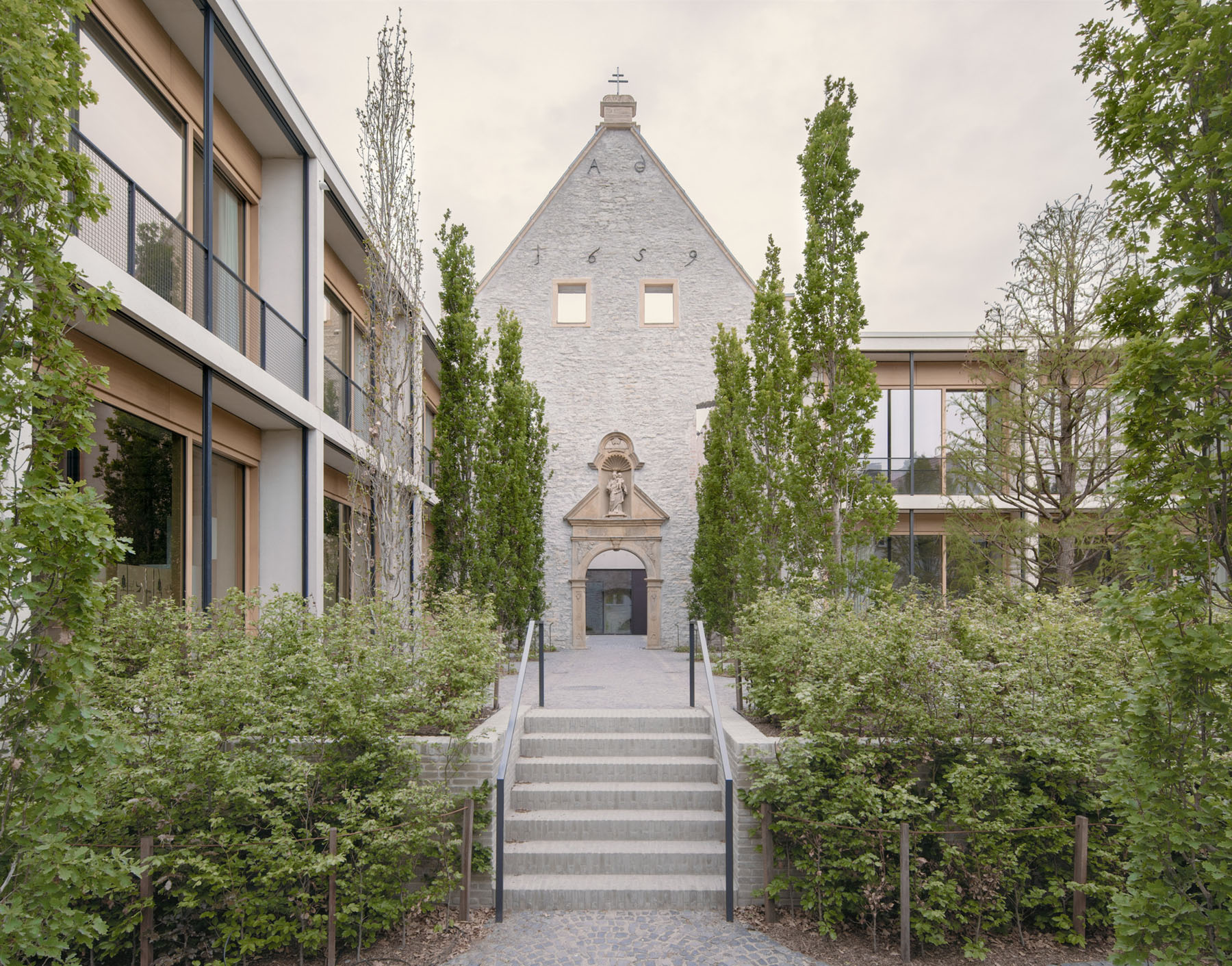 Jacoby Studios Headquarters by David Chipperfield Architects — A symmetrical axial view through a landscaped courtyard toward a converted historic chapel, its whitewashed brick facade punctuated by two square windows...
