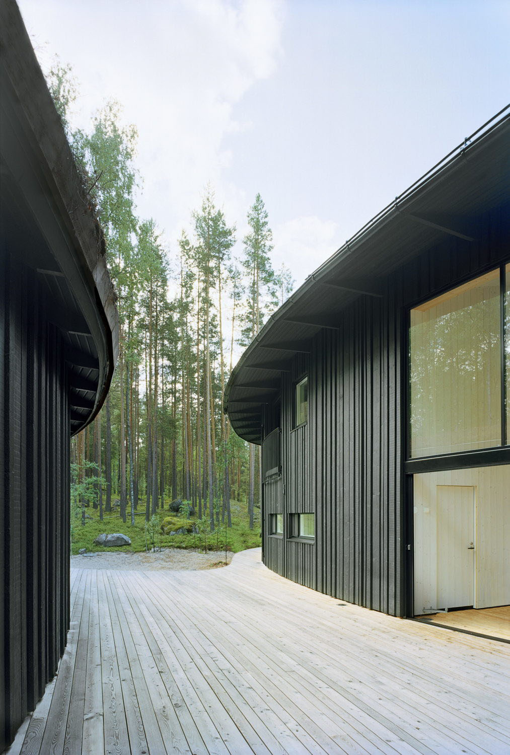 Villa Musu by Sanaksenaho Architects — A timber-clad passage frames the forest interior, where black-painted wooden facades in varied widths create visual rhythm along both sides of the wooden deck.
