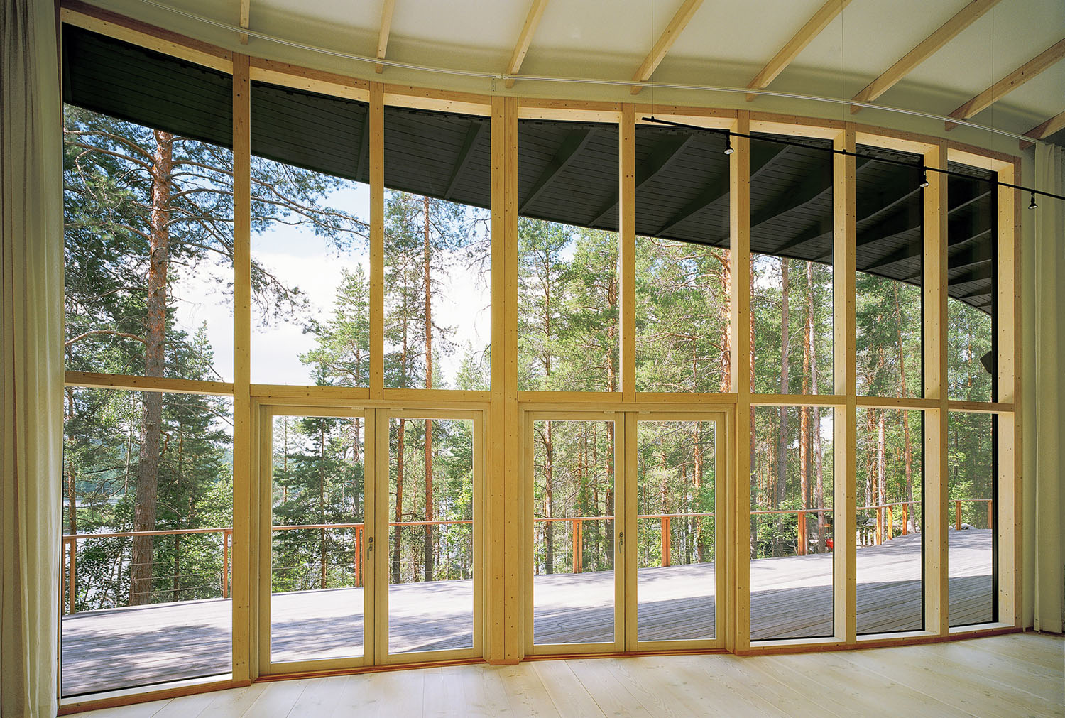 Villa Musu by Sanaksenaho Architects — Interior view of Villa Musu's south-facing glazed wall, a continuous expanse of timber-framed glass doors and windows opening onto a wooden deck.