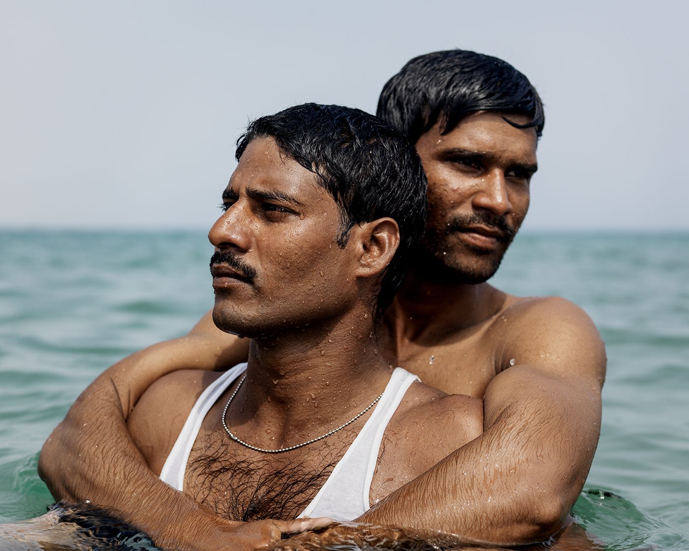 LIKE by Ryan Debolski — Two migrant workers stand in shallow seawater, their bodies wet from recent immersion.