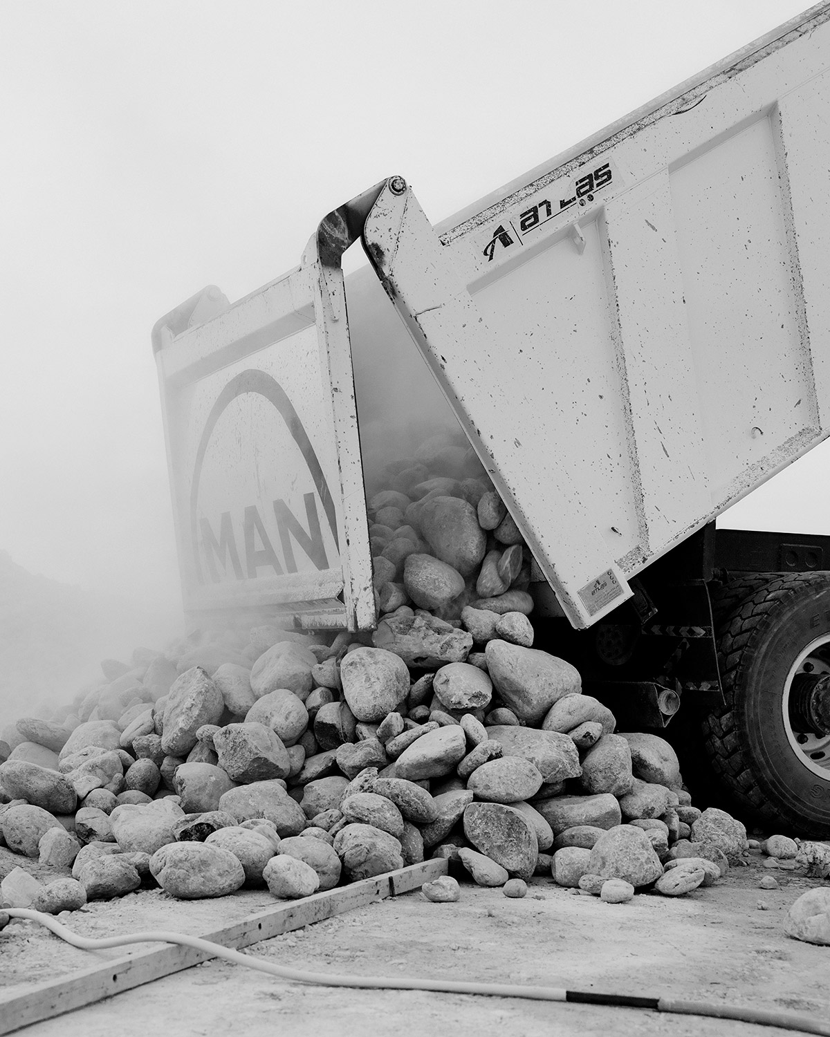 LIKE by Ryan Debolski — A MAN dump truck captured mid-discharge, its tilted bed releasing a substantial load of rounded aggregate stones onto unpaved ground.