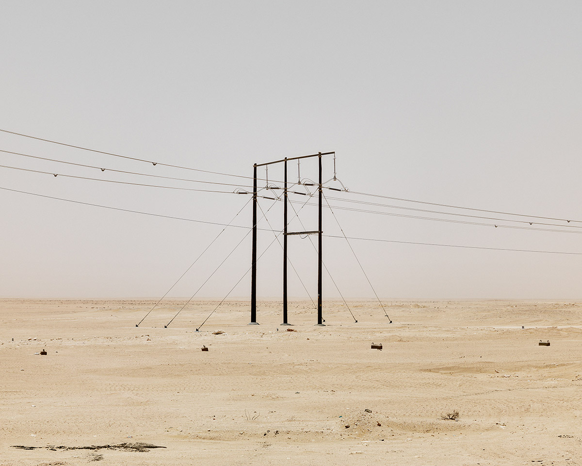 LIKE by Ryan Debolski — A steel transmission tower stands isolated in a vast desert landscape, its three vertical columns anchored by diagonal bracing cables and guy-wires...