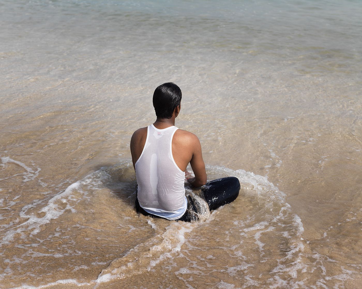 LIKE by Ryan Debolski — A man sits in shallow seawater wearing a white tank top, his back to the camera as waves lap around him.