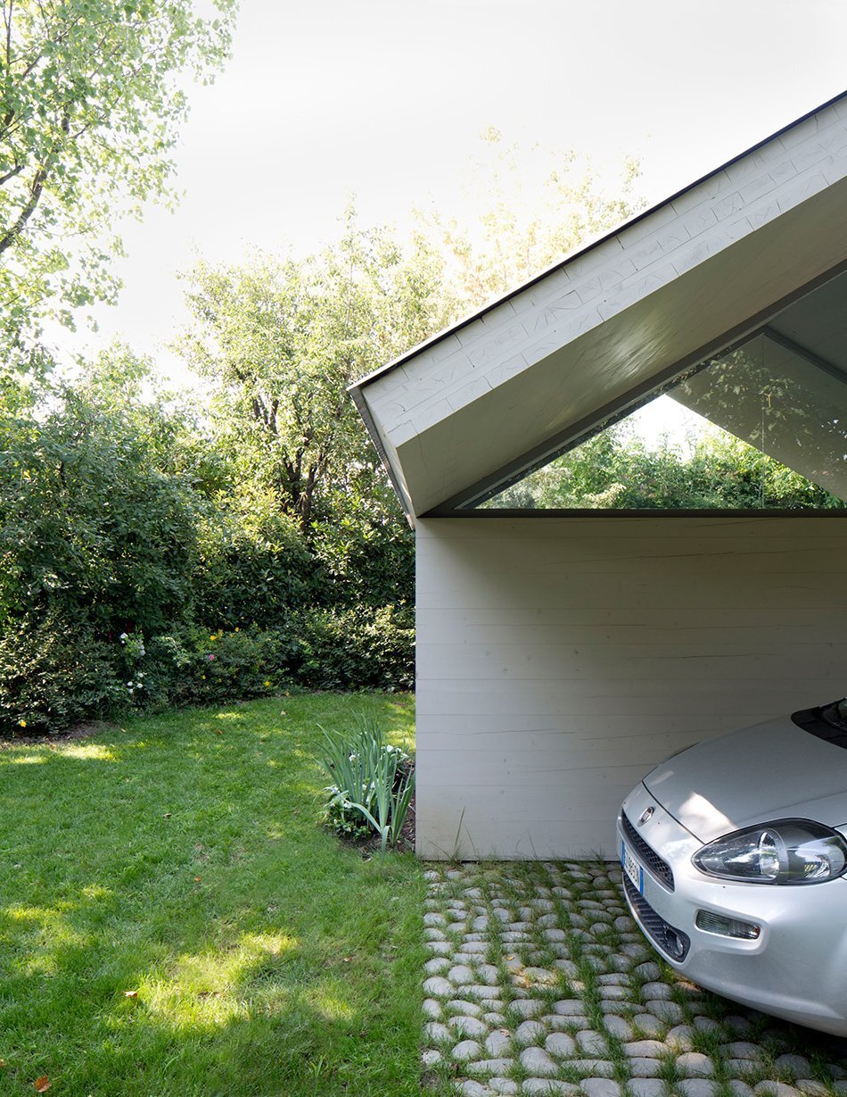 Garage and Workshop by Studiospazio — A white-rendered carport shelters a silver sedan beneath a corrugated metal gable roof with pronounced bilateral overhangs.