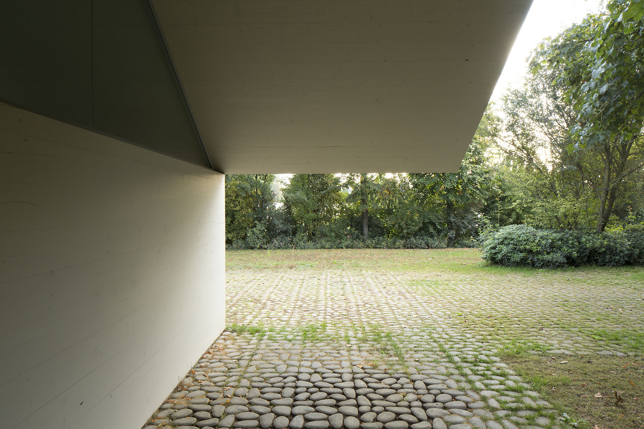 Garage and Workshop by Studiospazio — View from beneath the structure's overhang toward the paved courtyard and tree-lined perimeter.