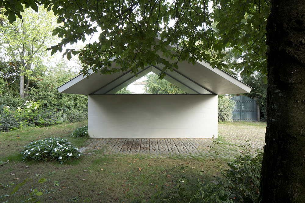 Garage and Workshop by Studiospazio — A cream-rendered pavilion with a pronounced pitched roof sits nestled within a mature woodland setting.