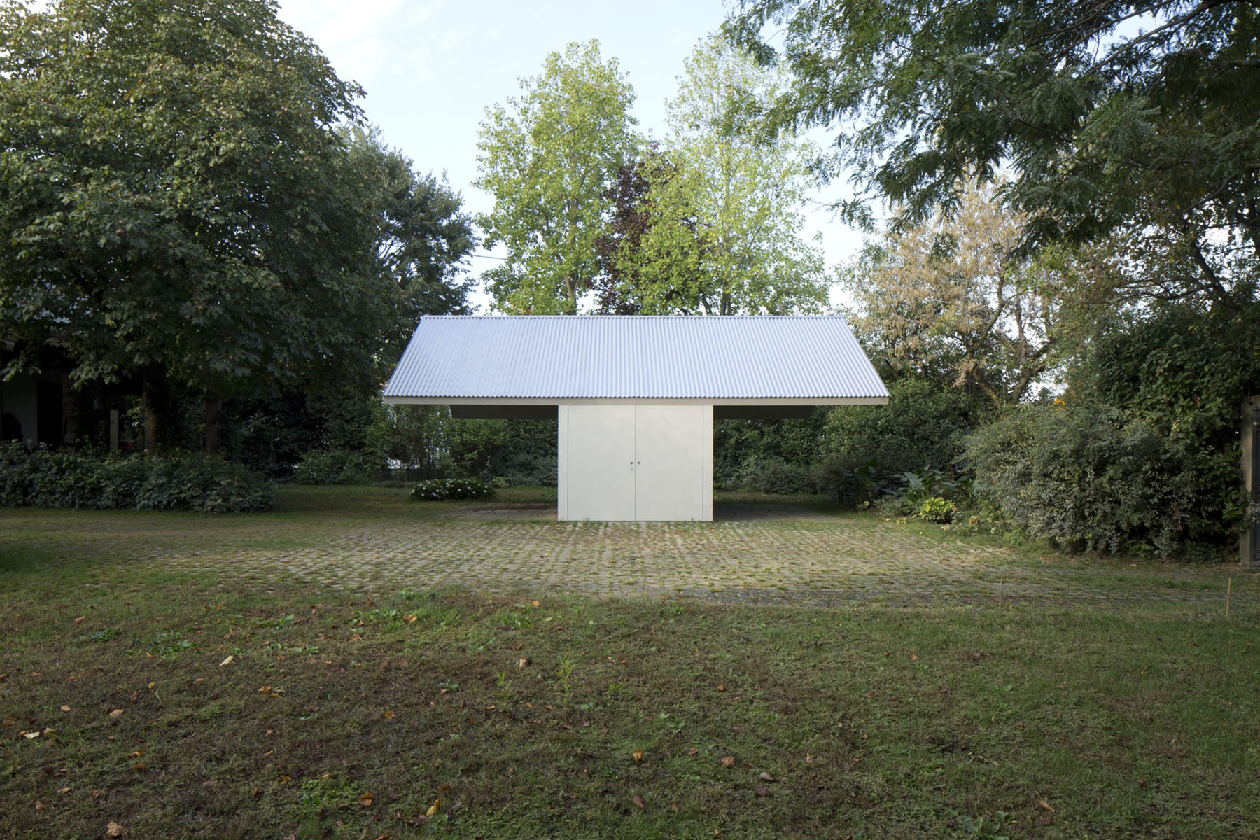 Garage and Workshop by Studiospazio — A modest pavilion structure stands isolated in a mowed clearing, surrounded by mature trees at various stages of seasonal change.