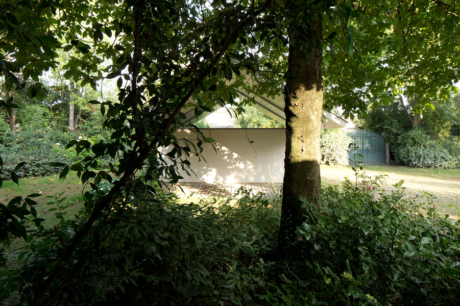 Garage and Workshop by Studiospazio — Framed through dense deciduous foliage, the workshop reveals its cream-rendered central volume beneath a timber gable roof with pronounced bilateral overhang.
