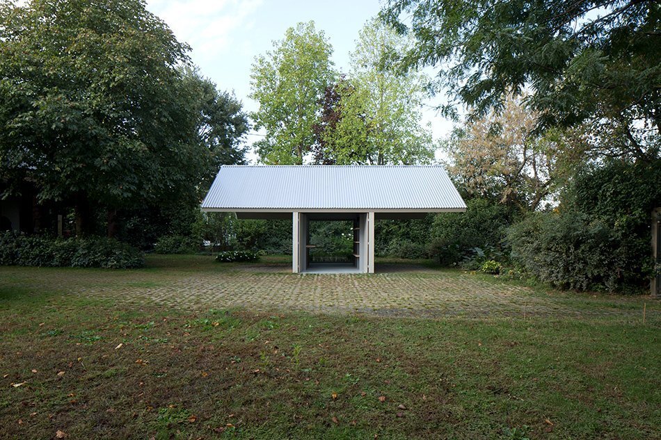 Garage and Workshop by Studiospazio — A modest pavilion structure set within a wooded landscape, anchored by a corrugated metal gable roof that extends generously on both sides.