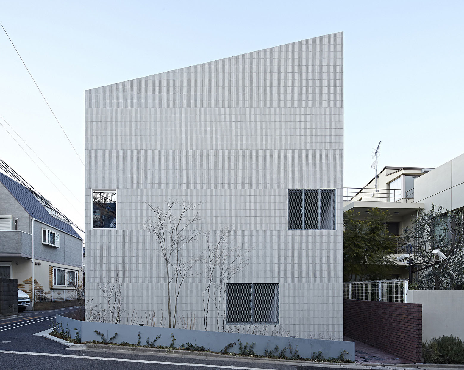Oggi by Makoto Yamaguchi Design — Oggi House presents a white ceramic tile facade organized in a rigid grid pattern, interrupted by three asymmetrically positioned dark-framed rectangular...