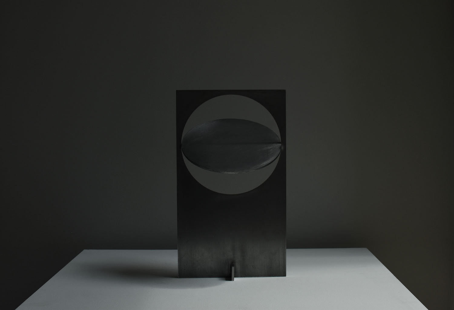 OBJ-01 by Manu Bañó — OBJ-01 presents a rotatable metal disc with a horizontal slot bisecting its circular form, mounted within a dark rectangular steel panel.