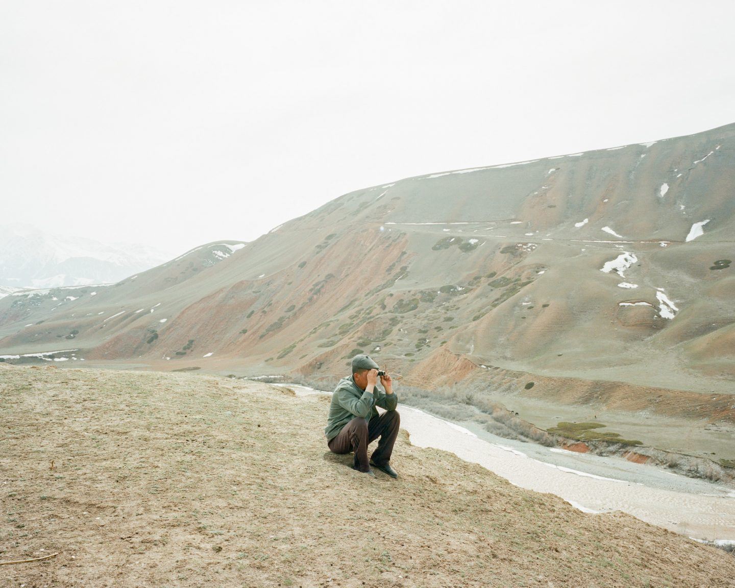 Horsehead Nebula by Matthieu Litt — A man in olive-green jacket and cap crouches on gravelly terrain, binoculars raised to his eyes, surveying a vast mountainous landscape.